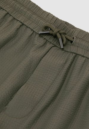Olívazöld, texturált szövetnadrág rugalmas derékpánttal és fém végződésű, megkötött húzózsinórral.