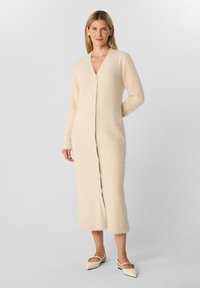 Cardigan lungo di colore crema realizzato in tessuto morbido e testurizzato, con scollatura a V, maniche lunghe e bottoni lungo la parte frontale fino a metà polpaccio.