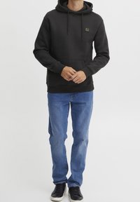 Felpa nera con cappuccio, con lacci, tasca a marsupio frontale e patch con logo. Indossata con jeans blu e scarpe da ginnastica nere.