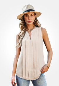 Beige mouwloze top met verticale plooien, V-hals en ronde zoom; gecombineerd met casual denim. Een gouden kettingaccentueert de look.