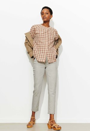 Femme debout contre un mur blanc, portant une chemise à carreaux boutonnée, une veste beige posée sur les épaules, un pantalon gris clair et des sandales compensées couleur fauve.