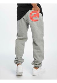 Grijze sweatpants met een relaxte pasvorm, voorzien van een oranje cirkelvormig logo op de achterzak. De stof is zacht met elastische boorden.