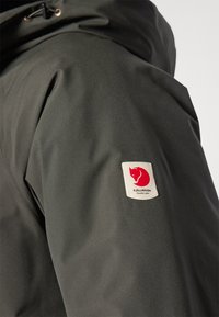 Donkergroene jas met een gladde, matte textuur. Voorzien van een rode vossensymbool op de bovenarm en een verstelbare trekkoord in de capuchon.