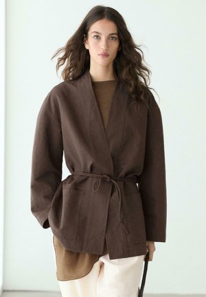 Frau mit welligem braunem Haar trägt eine dunkelbraune Jacke mit Gürtel über einem braunen Oberteil und hellbeigen Hosen, stehend vor hellem Hintergrund.