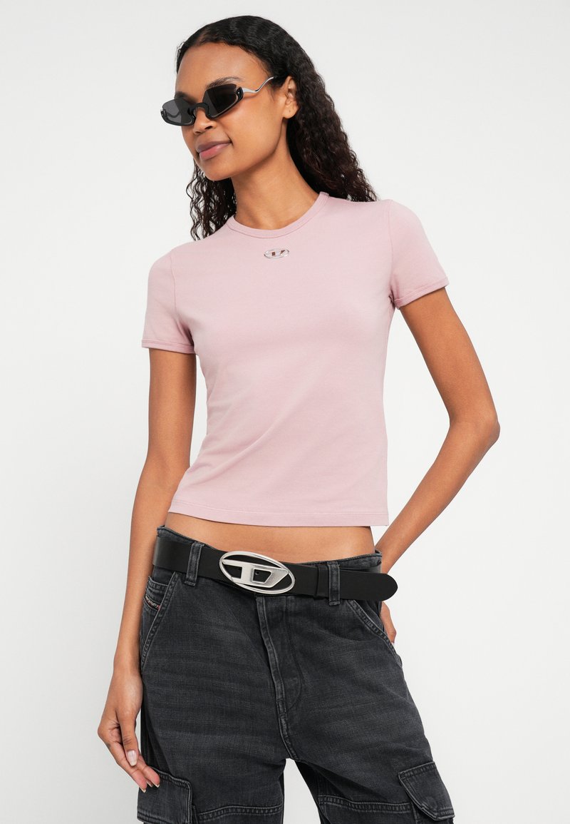 Lyserød tætsiddende t-shirt med korte ærmer, sølvfarvet logodetalje på brystet; kombineret med højtaljede sorte cargobukser og et markant bælte.