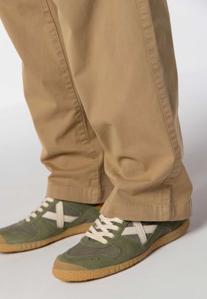 Sneakers verdi e grigie con dettagli bianchi e lacci beige, abbinate a pantaloni beige larghi che mostrano un design casual e strutturato.