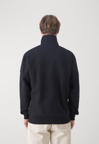 Sudadera negra con cuello alto, puños y dobladillo acanalados, hecha de un tejido suave. Detalle de costura visible en la parte posterior.