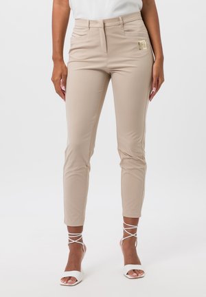 Pantalons slim beige en tissu extensible avec poches avant. Accentue par un petit logo métallique à l'avant, associés à des talons blancs à lanières.