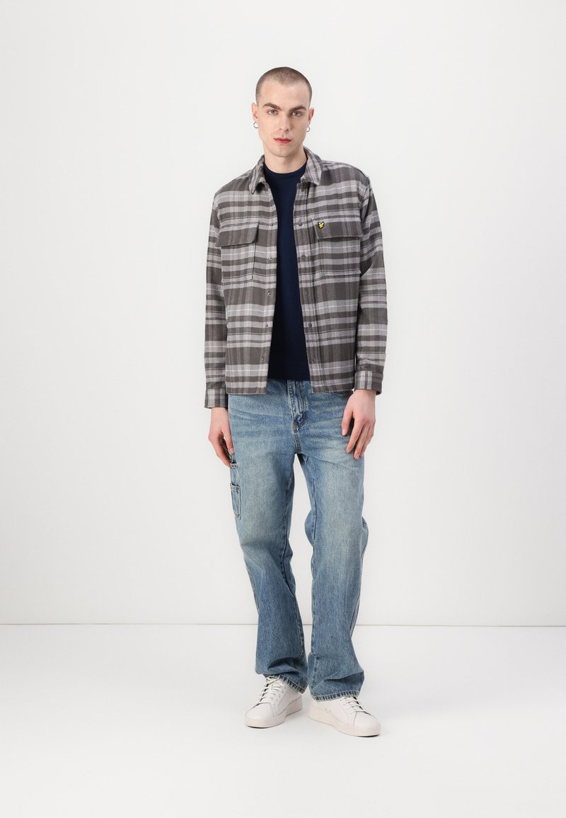 Grå rutete flanellskjorte med to brystlommer, mørkeblå t-skjorte under, kombinert med lyseblå baggy jeans og hvite joggesko.