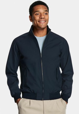 Marineblaue leichte Jacke mit Stehkragen, Reißverschluss vorne, Seitentaschen und gerippten Bündchen, kombiniert mit einem hellblauen Hemd darunter.