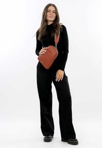 Bolso cruzado de cuero marrón con forma rectangular, superficie texturada, cierre superior con cremallera y correa ajustable. Modelo lleva un atuendo negro.