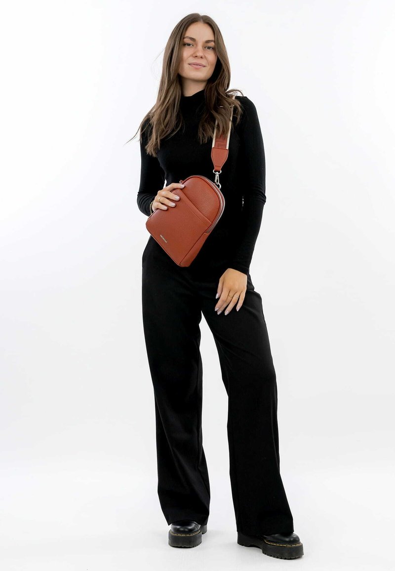 Bolso cruzado de cuero marrón con forma rectangular, superficie texturada, cierre superior con cremallera y correa ajustable. Modelo lleva un atuendo negro.