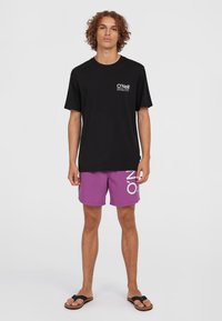 Melns īsām piedurknēm t-krekls ar baltu logo, savienots ar purpura peldētšortiem, kurus rotā liels balts "O" prints, papildināts ar melnām čības.