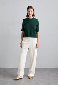 Maglione verde a maniche corte lavorato a maglia con orlo a coste, abbinato a pantaloni bianchi a gamba larga. Il modello indossa mocassini dorati con dettaglio a fiocco.