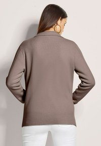 Braune Strickjacke mit lockerer Passform, langen Ärmeln und Schalkragen. Sie hat einen strukturierten Stoff und wird über weißen Hosen getragen.