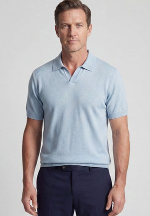 Man die een lichtblauw poloshirt met korte mouwen en donkerblauwe broek draagt, staand tegen een effen lichte achtergrond.