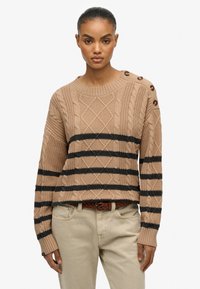 Gebreide trui in lichtbruin met zwarte strepen. Kenmerkt zich door textuurpatronen, een ronde halslijn en knopen op de schouder. Gepaard met beige broek.