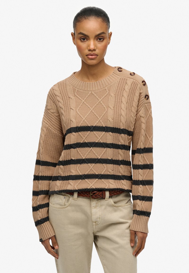 Gebreide trui in lichtbruin met zwarte strepen. Kenmerkt zich door textuurpatronen, een ronde halslijn en knopen op de schouder. Gepaard met beige broek.