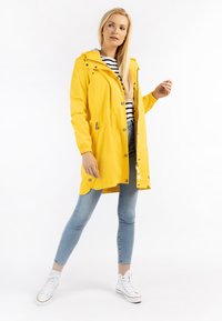 Impermeabile giallo con cappuccio regolabile, chiusura a zip e bottoni a pressione, e tasche laterali. Indossato sopra una maglietta a righe, abbinato a jeans azzurri e sneakers bianche.