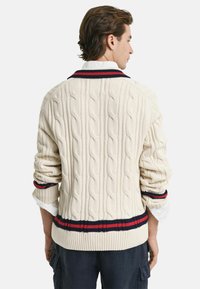 Kabelstrikket cremefarvet sweater med ribkant forneden og ved ærmerne. Har marineblå og røde striber på kraven og ærmerne. Casual design med struktureret stof.