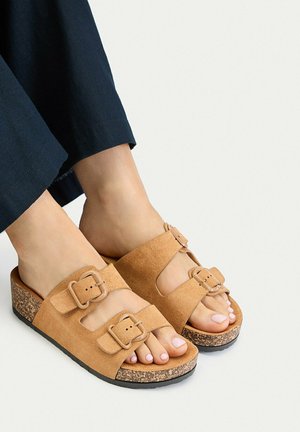 Pies con sandalias de ante marrón claro con dos tiras ajustables con hebilla y suelas de cuña de corcho, combinadas con pantalones oscuros recortados.