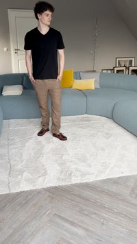 Schwarzes V-Ausschnitt-T-Shirt und beigefarbene Hose, getragen mit braunen Bootsschuhen. Gebogene blaue Couch, weißer strukturierter Teppich und helle Holzfußböden.