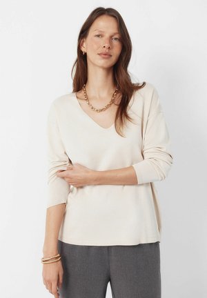 Beżowy sweter z dekoltem w serek, długie rękawy, miękka faktura i luźny krój. Noszony z szarymi spodniami i nałożoną warstwowo złotą biżuterią.