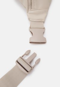 Béžový nastavitelný popruh s opalovacím plastovým přezkou a sponou; obsahuje texturovaný nylonový pás a hladkou látku poblíž zipu.