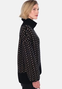 Tjock svart tröja med hög krage, med ett kontrasterande beige mönster. Designen inkluderar geometriska former och strukturerad stickning.