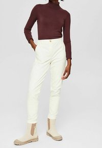 Pantaloni a vita alta color crema con un bottone dorato, abbinati a un dolcevita aderente bordeaux e stivaletti beige con pannelli laterali.