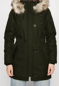 Parka verde con capucha de piel desmontable, broches a presión, bolsillos laterales, cintura elástica y textura suave.