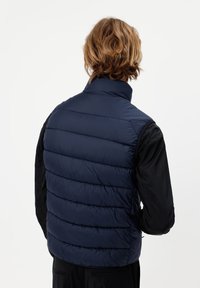 Osoba o średniej długości jasnobrązowych włosach, ubrana w granatową pikowaną kamizelkę na czarną koszulkę z długim rękawem, widoczna od tyłu.