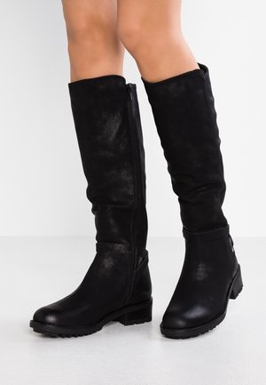 Bottes noires montantes jusqu'aux genoux avec de petits talons blocs portées par une personne debout sur une surface blanche devant un fond uni.