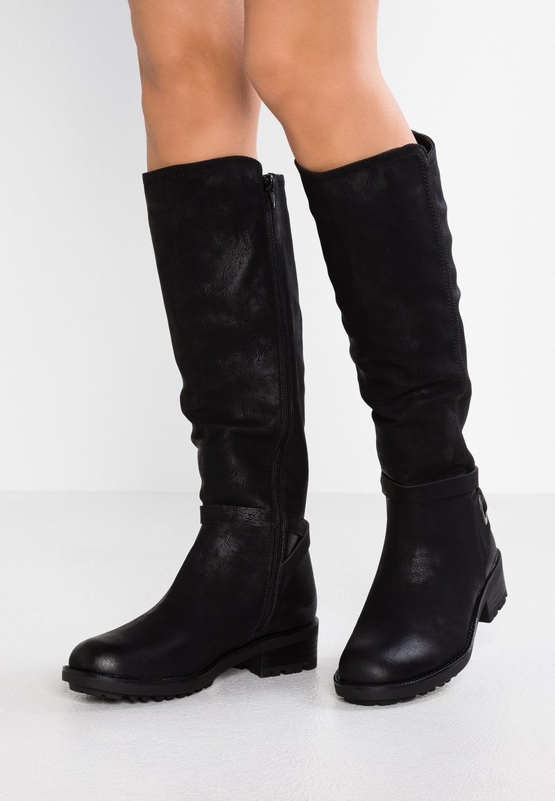 Bottes noires montantes jusqu'aux genoux avec de petits talons blocs portées par une personne debout sur une surface blanche devant un fond uni.