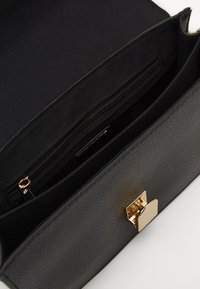Bolso de mano negro texturizado con un cierre dorado; cuenta con dos compartimentos, uno con cremallera y un interior de tela suave.