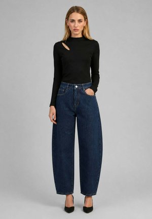 Femme portant un haut noir à manches longues avec une découpe et un jean bleu foncé taille haute et jambes larges avec des talons noirs.