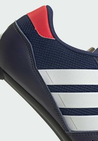 Zapatilla deportiva azul con parte superior de malla y detalles en rojo, tres rayas blancas y un acabado de cuero texturizado. Suela de goma duradera.