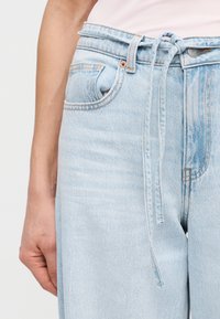 Detailný záber na svetlomodré riflové nohavice s uviazaným látkovým opaskom a viditeľnou vreckovou časťou, ktoré nosí osoba v ružovom topu.