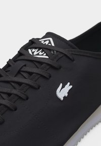 Μαύρο δερμάτινο αθλητικό παπούτσι με λευκό λογότυπο κροκοδείλου στο πλάι και τριγωνικό λογότυπο στη γλώσσα, σε λευκό φόντο.