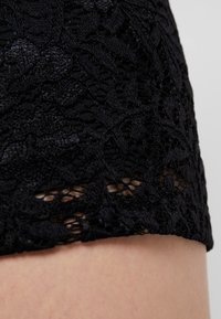Černá krajková tkanina s jemnými květinovými vzory, strukturovaným povrchem a lemováním ve tvaru klobouku. Detailní záběr ukazuje jemné detaily a otevřenost.