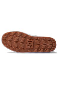 Outsole skate topánky DC s hnedým gumovým vzorom, s textúrovaným povrchom a logom v strede. Biele okraje obrysujú okraje.