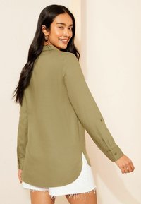 Camicia a maniche lunghe verde oliva in tessuto leggero, con polsini con bottoni e orlo curvato, abbinata a pantaloncini bianchi sfrangiati.