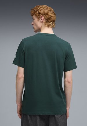 Joven con cabello rizado rojo que lleva una camiseta de manga corta verde oscuro y pantalones gris a cuadros, de espaldas a la cámara.