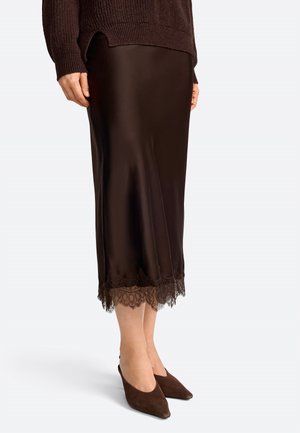 Dunkelbrauner Satinrock mit einer Spitzenborte am Saum und geradem Schnitt. Kombiniert mit spitzen, braunen Wildlederschuhen.