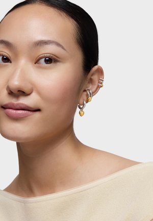 Młoda kobieta o gładkiej skórze, nosząca wiele srebrnych i złotych kolczyków kołowych, w tym z zawieszką w kształcie serca, na jednolitym jasnym tle.