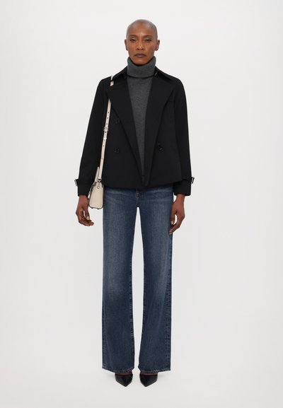 Manteau noir à double boutonnage sur un col roulé gris, associé à un jean bleu foncé évasé et des talons noirs à bout pointu. Petit sac à main beige.
