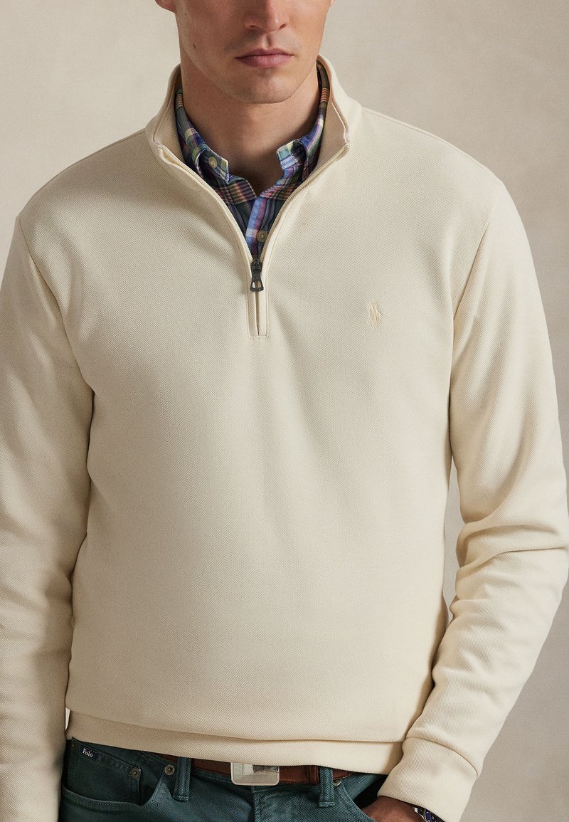 Man wearing cream-colored quarter-zip sweater over blue, green, and purple plaid button-up shirt, with teal pants and brown belt.