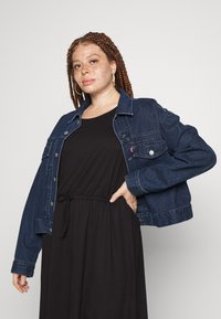Casaco de ganga com lavagem escura, fechos de botão, dois bolsos frontais e um corte descontraído, combinado com um vestido preto com cintura ajustável por cordão.