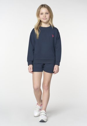 Ragazza giovane con lunghi capelli biondi che indossa una felpa blu navy, pantaloncini abbinati e scarpe da ginnastica bianche con velcro, che cammina in avanti su uno sfondo neutro.