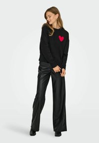 Maglione nero con una trama a cuore rosso sul petto, abbinato a pantaloni in ecopelle nera lucida a vita alta e scarpe nere.